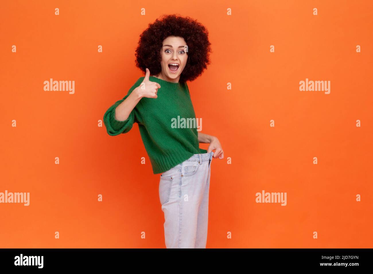 Portrait of positive excited woman with Afro hairstyle wearing green casual style sweater showing slim waist in big trousers and thumbs up. Indoor studio shot isolated on orange background. Stock Photo