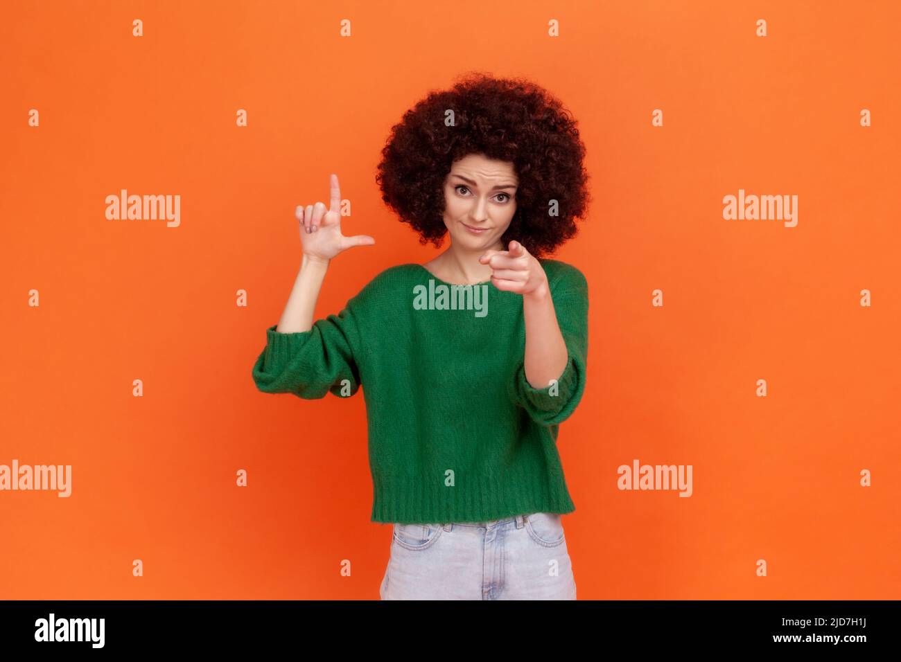 You are loser. Woman with Afro hairstyle wearing green casual style sweater showing loser gesture and pointing finger on you, abuser. Indoor studio shot isolated on orange background. Stock Photo