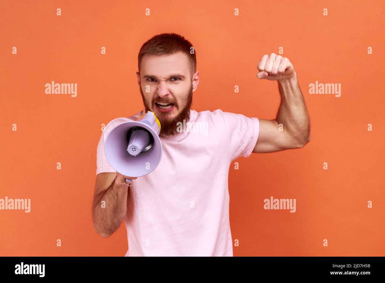 Portrait of angry bearded man loudly screaming at megaphone, making announce, protesting, wants to be heard, wearing pink T-shirt. Indoor studio shot isolated on orange background. Stock Photo