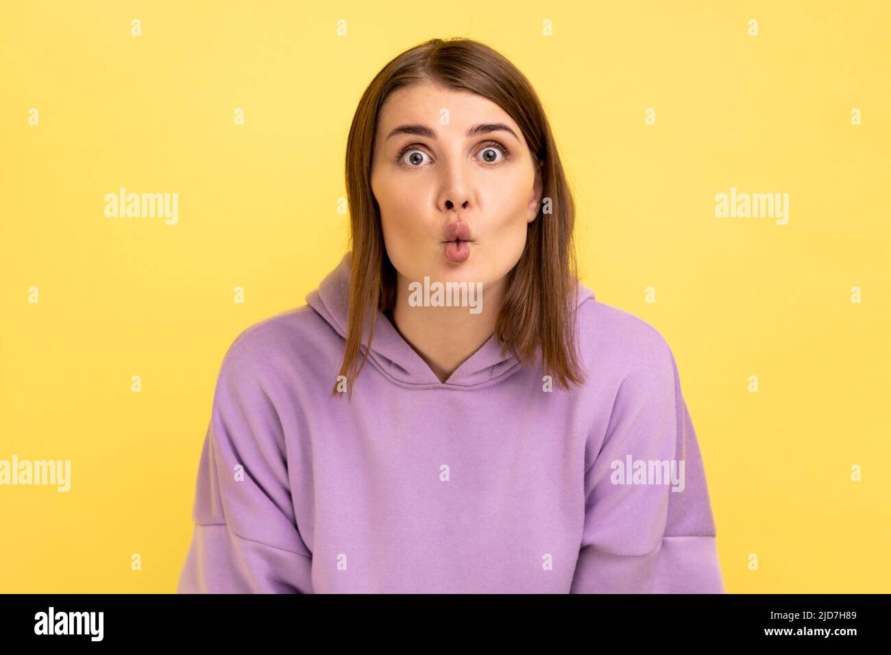 Portrait of cute funny woman expressing amazement with fish face grimace, pout lips and big eyes, looking shocked and comical, wearing purple hoodie. Indoor studio shot isolated on yellow background. Stock Photo