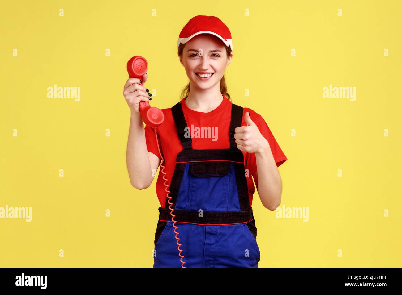 Portrait of delighted worker woman holding out handset, showing thumb up, likes order service, looking friendly at camera, wearing overalls and cap. Indoor studio shot isolated on yellow background. Stock Photo