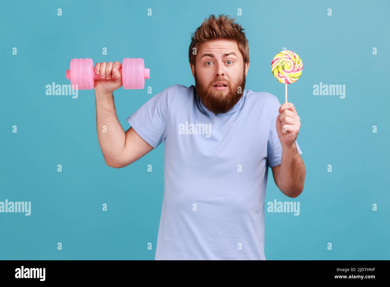 Portrait of puzzled confused handsome bearded man holding lollypop in hand and having training with pink dumbbell, shrugging shoulders. Indoor studio shot isolated on blue background. Stock Photo