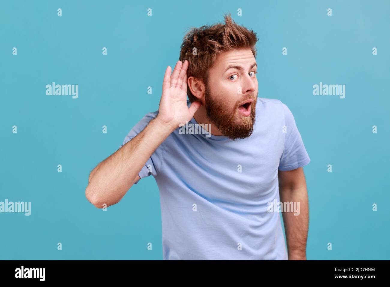 Can't hear you. Portrait of attentive bearded man holding hand near ear trying to listen quiet conversation, overhearing gossip, having hearing problems. Indoor studio shot isolated on blue background Stock Photo