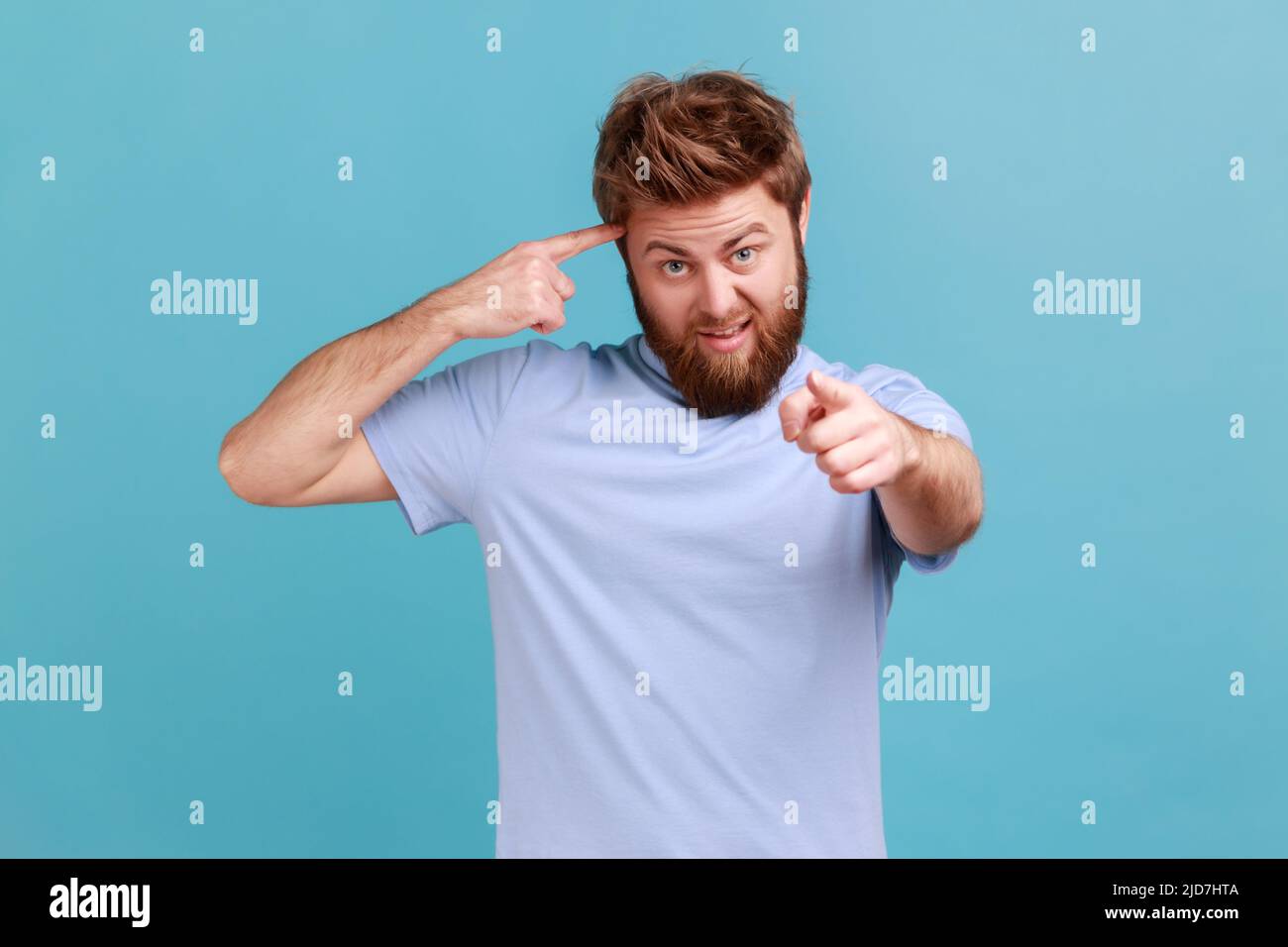 Hey, you are kidding. Portrait of bearded man with surprised face holding finger near head and pointing finger at camera, stupid gesture. Indoor studio shot isolated on blue background. Stock Photo