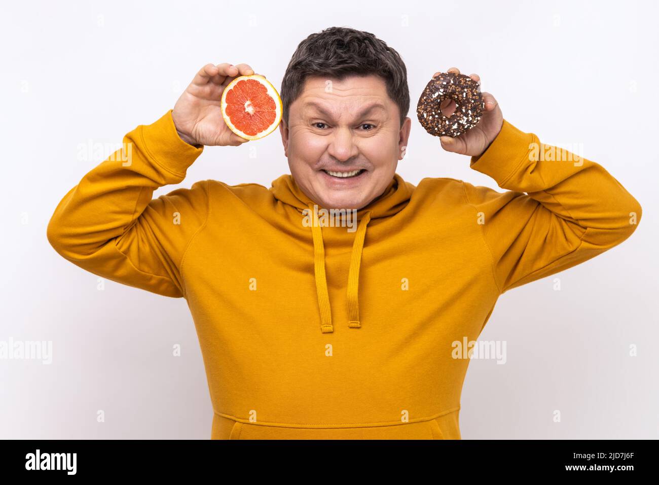 Portrait of positive funny man showing sweet doughnut and fresh juicy grapefruit, carbs and glucose in diet, wearing urban style hoodie. Indoor studio shot isolated on white background. Stock Photo