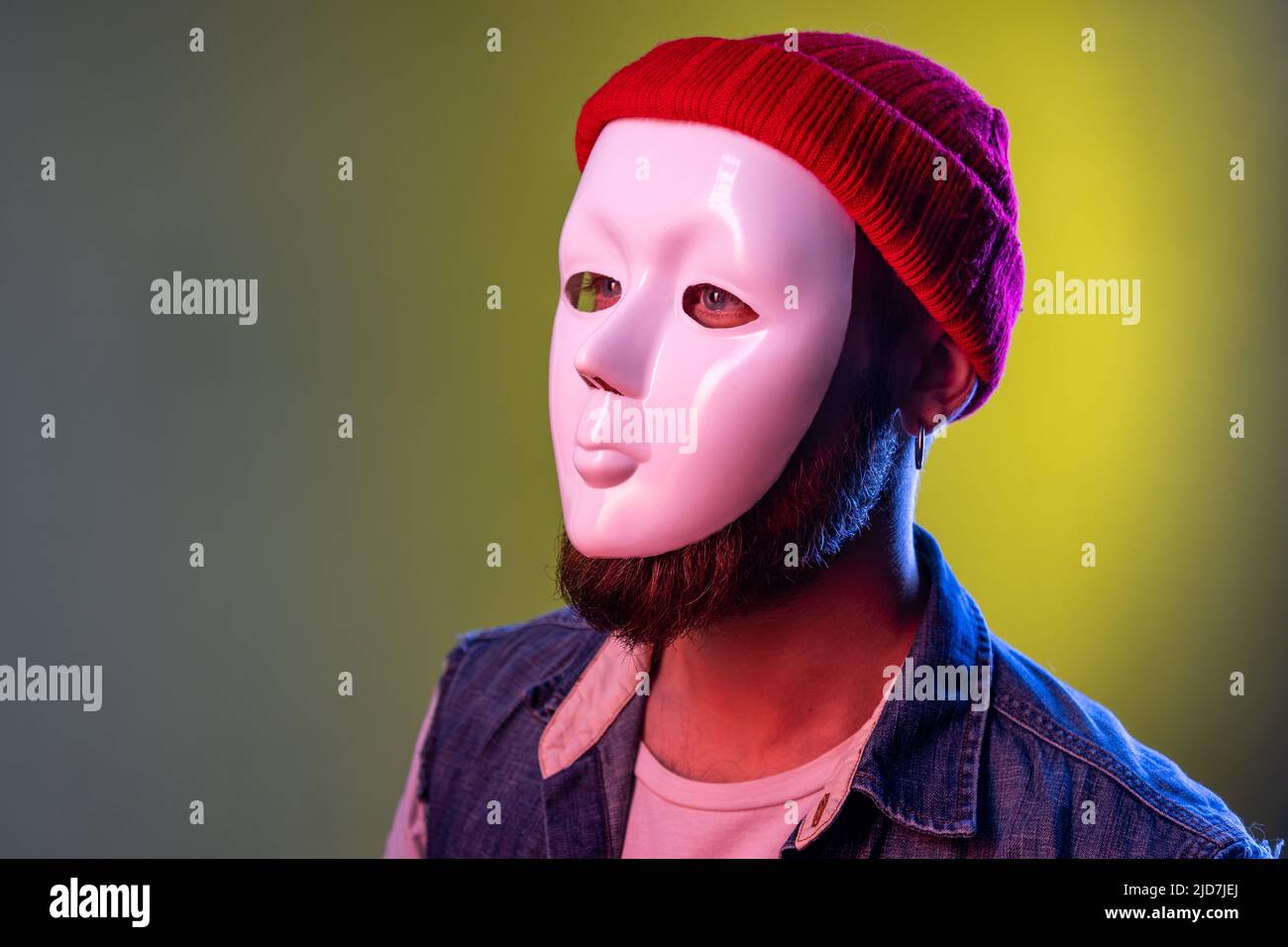 Side view of anonymous hipster man in white mask on face, hiding his identity, spying undercover, corruption, wearing hat and denim vest. Indoor studio shot isolated on colorful neon light background. Stock Photo