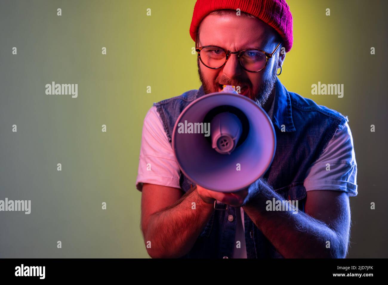 Angry hipster man holding megaphone near mouth loudly speaking, screaming, making announcement, paying attention at social problems. Indoor studio shot isolated on colorful neon light background. Stock Photo
