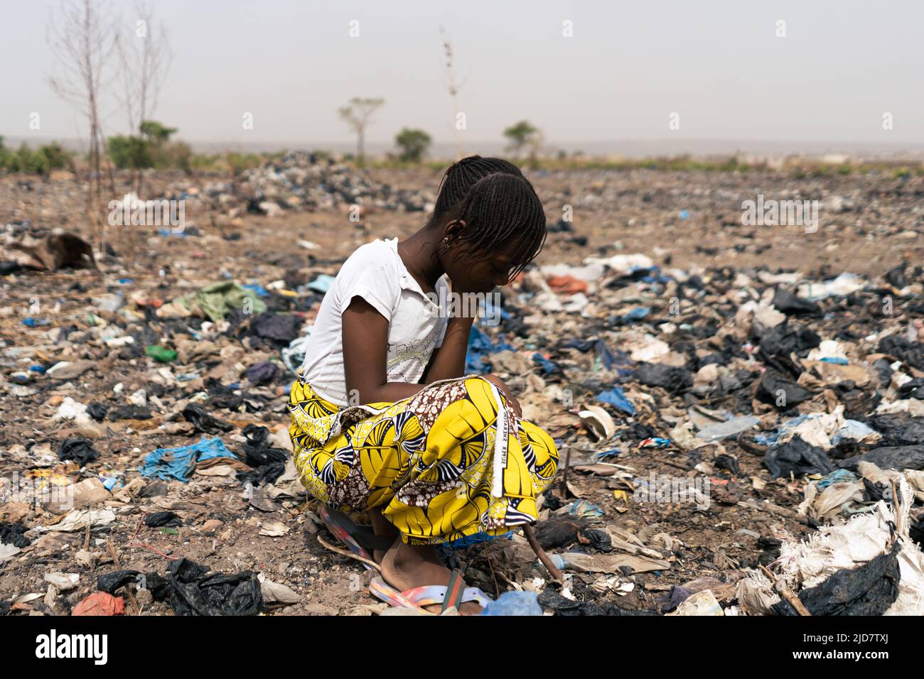 Exhausted little African girl kneeling in a garbage dump where she is forced to search for reusable material; child labor and exploitation Stock Photo