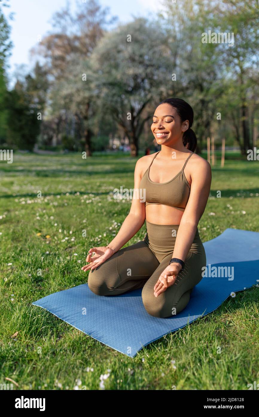 Peaceful millennial black lady sitting on sports mat, meditating with closed eyes at urban park, copy space Stock Photo