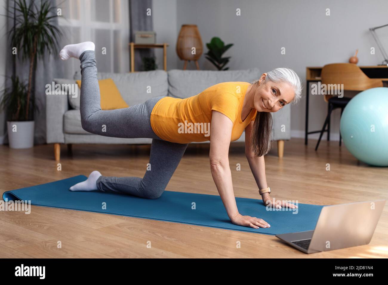 Online home fitness. Senior woman exercising to sports video on laptop in living room, looking at camera and smiling Stock Photo