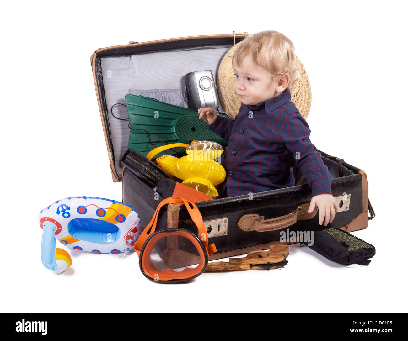 A little boy sits inside a suitcase with travel equipment, on white