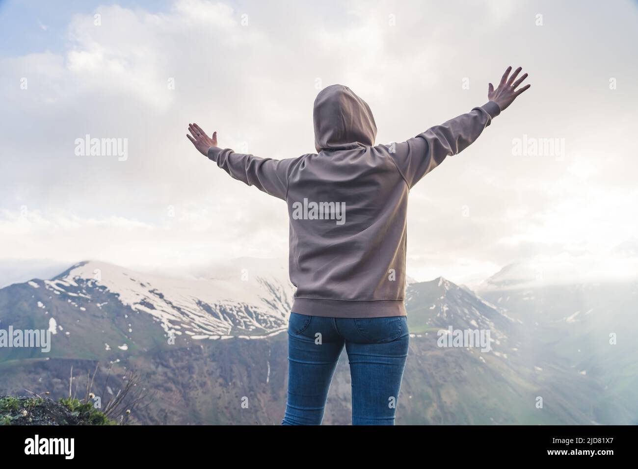 Travel concept. Woman dressed in a hoodie and jeans with her arms lifted up looking at a mesmerizing panorama of high mountain range. High quality photo Stock Photo