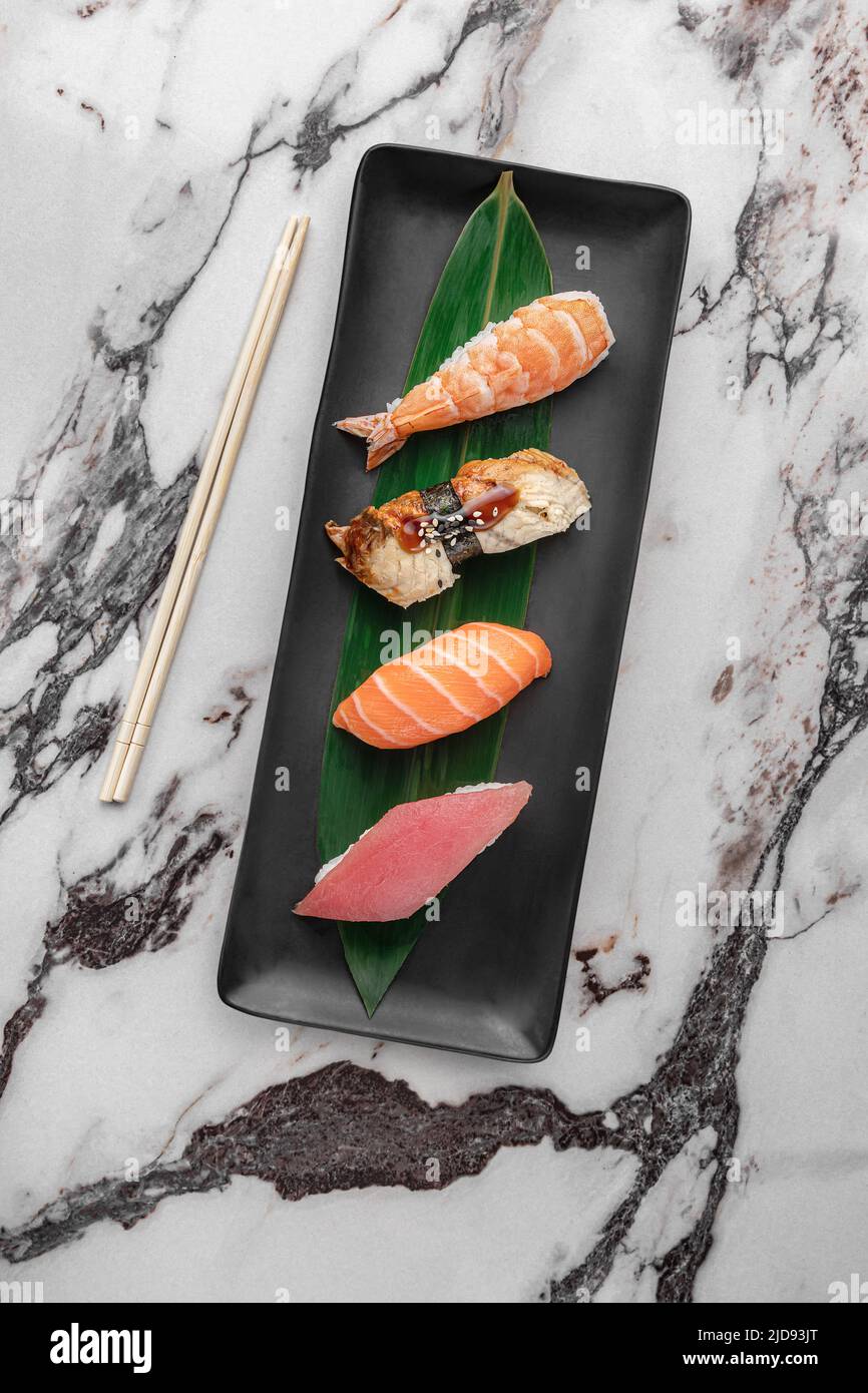 set of nigiri with tiger shrimp, tomago, salmon, tuna and green bamboo leaf in a black ceramic plate with chopstick on a bright white textured marble Stock Photo