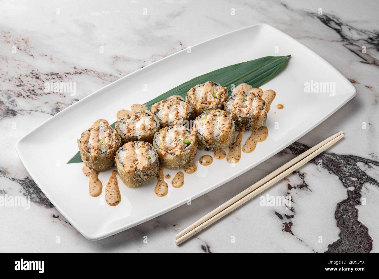 set of fried rolls with tiger shrimp, avocado, sesame sauce and green bamboo leaf in a white ceramic plate with chopstick on a bright textured marble Stock Photo