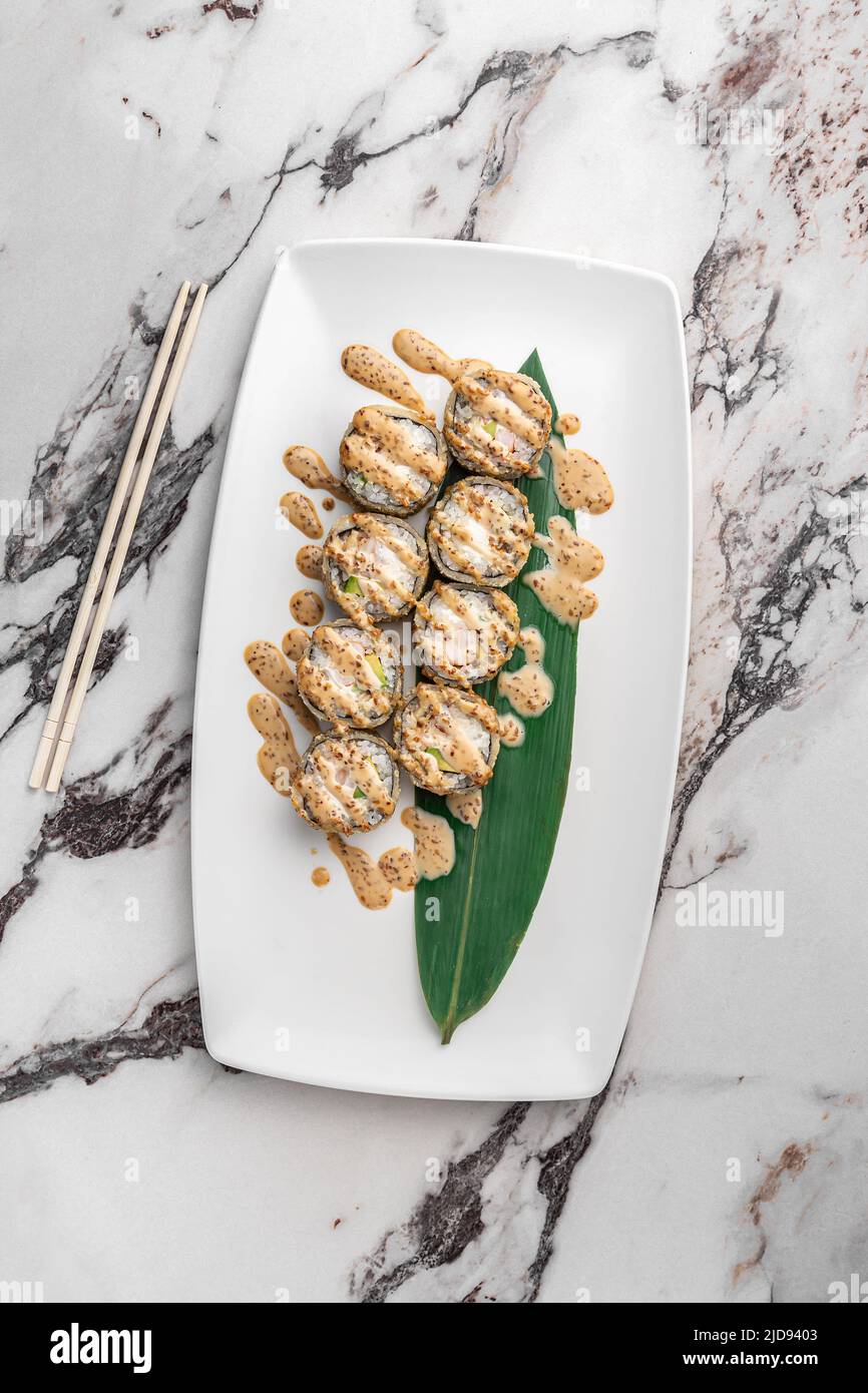 set of fried rolls with tiger shrimp, avocado, sesame sauce and green bamboo leaf in a white ceramic plate with chopstick on a bright textured marble Stock Photo