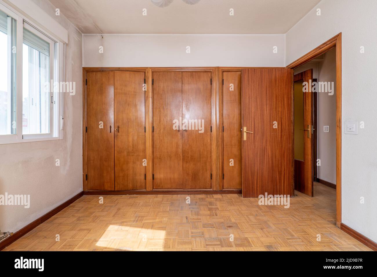 Room with a wall full of fitted wardrobes with six smooth wooden doors and double aluminum windows Stock Photo