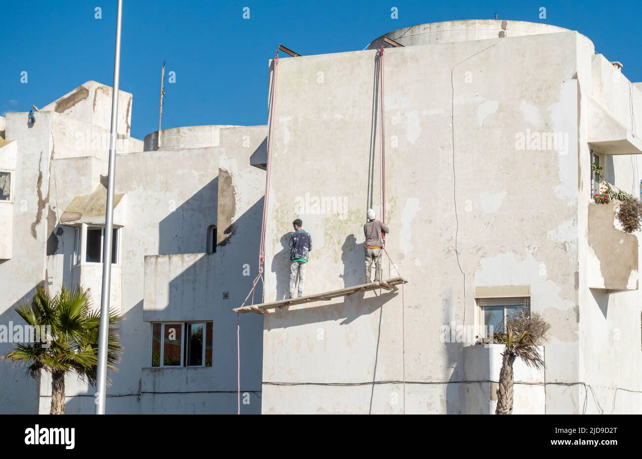 Workers on hanging platform painting remodelling building in  Lotissement Amal district in Essaouira, Morocco Stock Photo