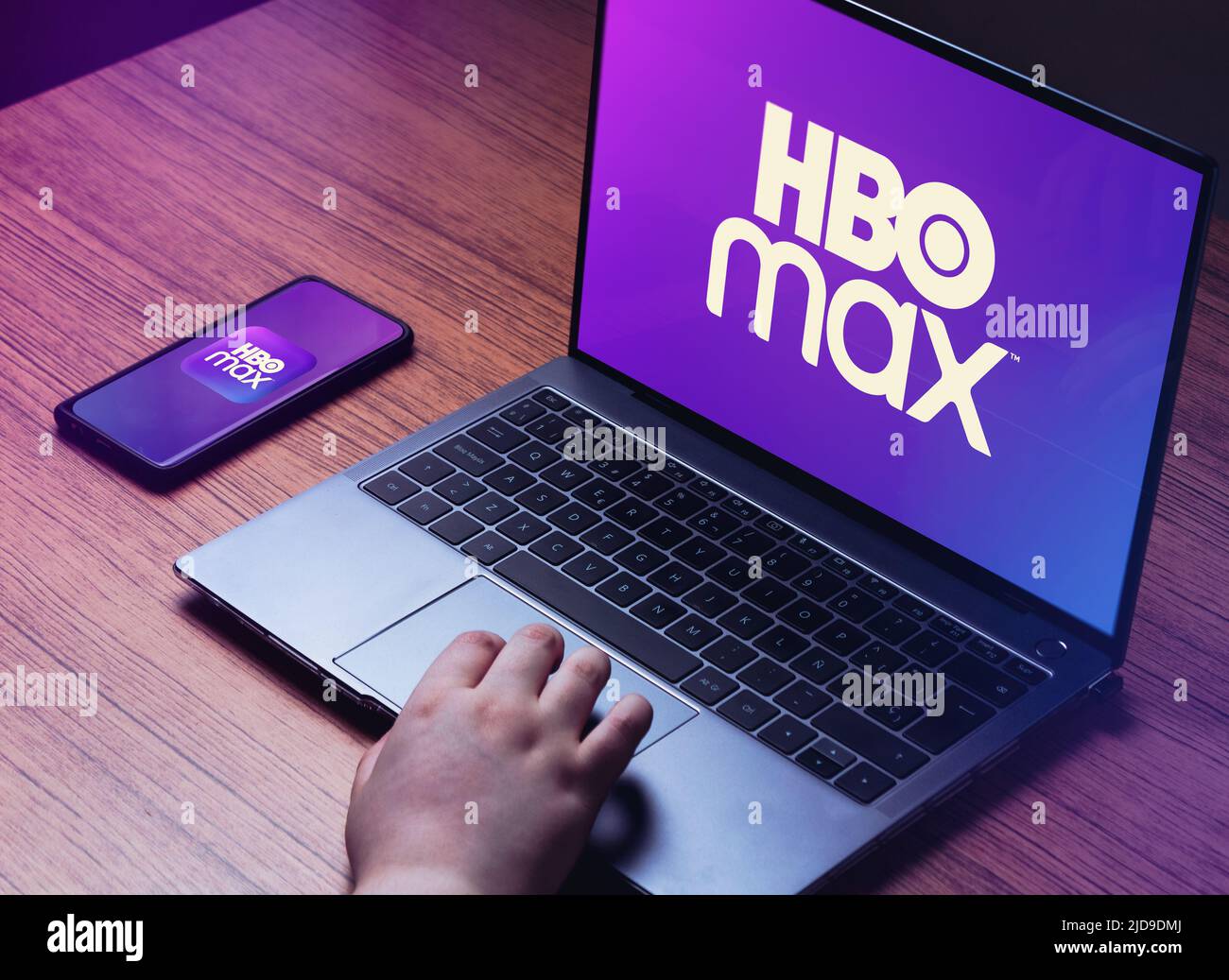 Young woman watching HBO Max on laptop. HBO Max logo on laptop and smartphone screens. Wood desk with laptop and smart Stock Photo