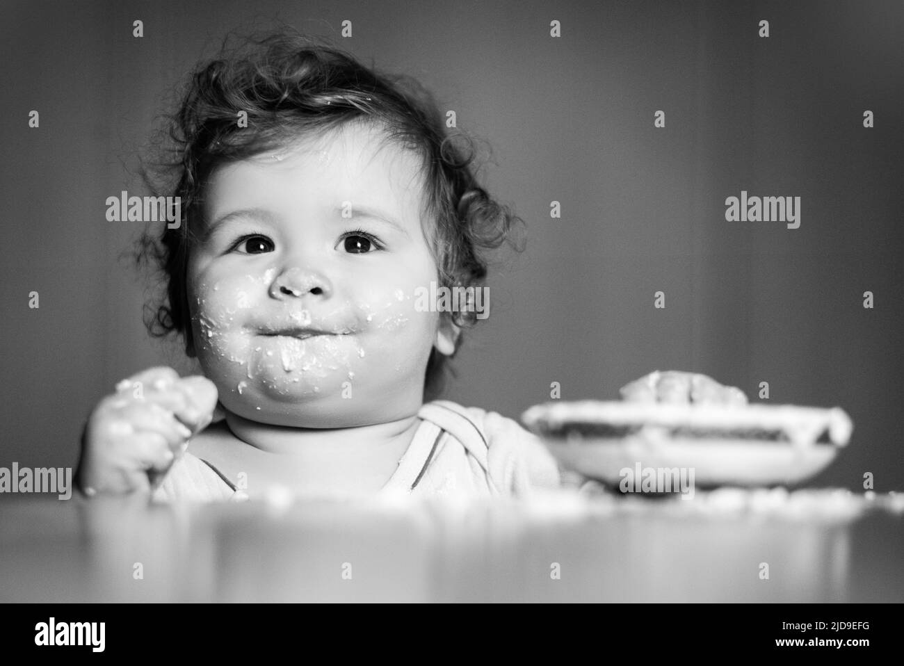 Laughing funny baby with dirty food face. Happy child spoon eats itself. Stock Photo