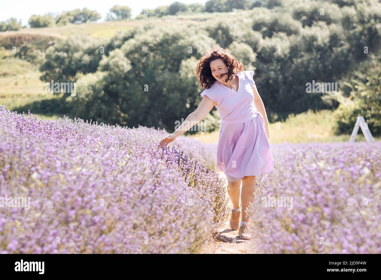 Soft selective focus girl in purple dress walks on lavender field and enjoys the aroma of pink flowers and beauty on sunny day. Trendy color of 2022 Very peri. Digital detox pleasure of slow life. Stock Photo