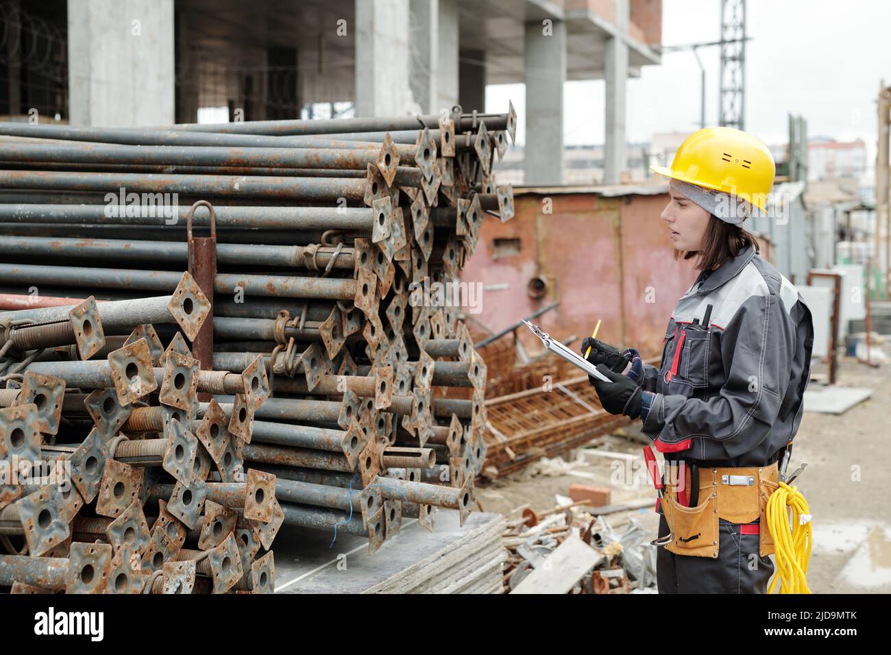 Young female engineer in workwear making notes in document while standing in front of two stacks of metallic pipes at construction area Stock Photo