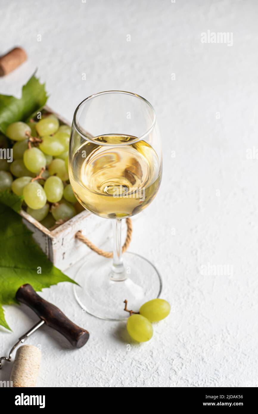 White wine glass still life with grapes in wooden box, corkscrew on white textured background. Text space right. Vertical orientation Stock Photo