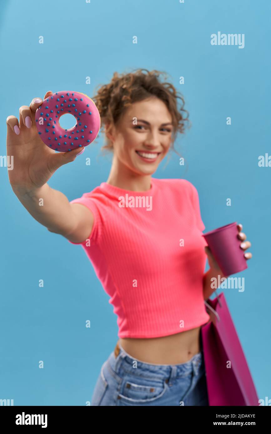 Happy lady holding pink donut, looking at camera indoor. Front view of cheerful blurred girl holding round donut in front of face, smiling, isolated on blue studio background. Concept of fast food. Stock Photo