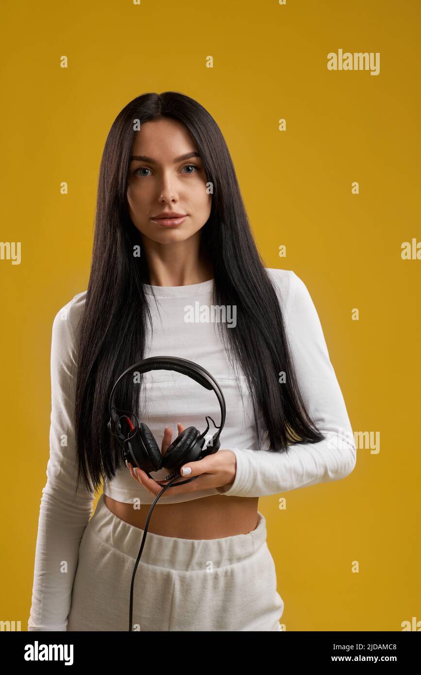 Attractive brunette girl in white shirt carefully holding headphones, while looking at camera. Portrait view of sporty woman with earphones, isolated on orange studio background. Concept of music. Stock Photo
