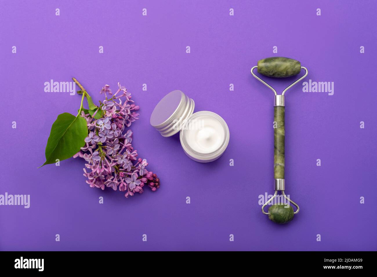 Jade facial roller, cream jar and lilac flower on purple background. Natural cosmetics, skin care concept. Top view, flat lay. Stock Photo