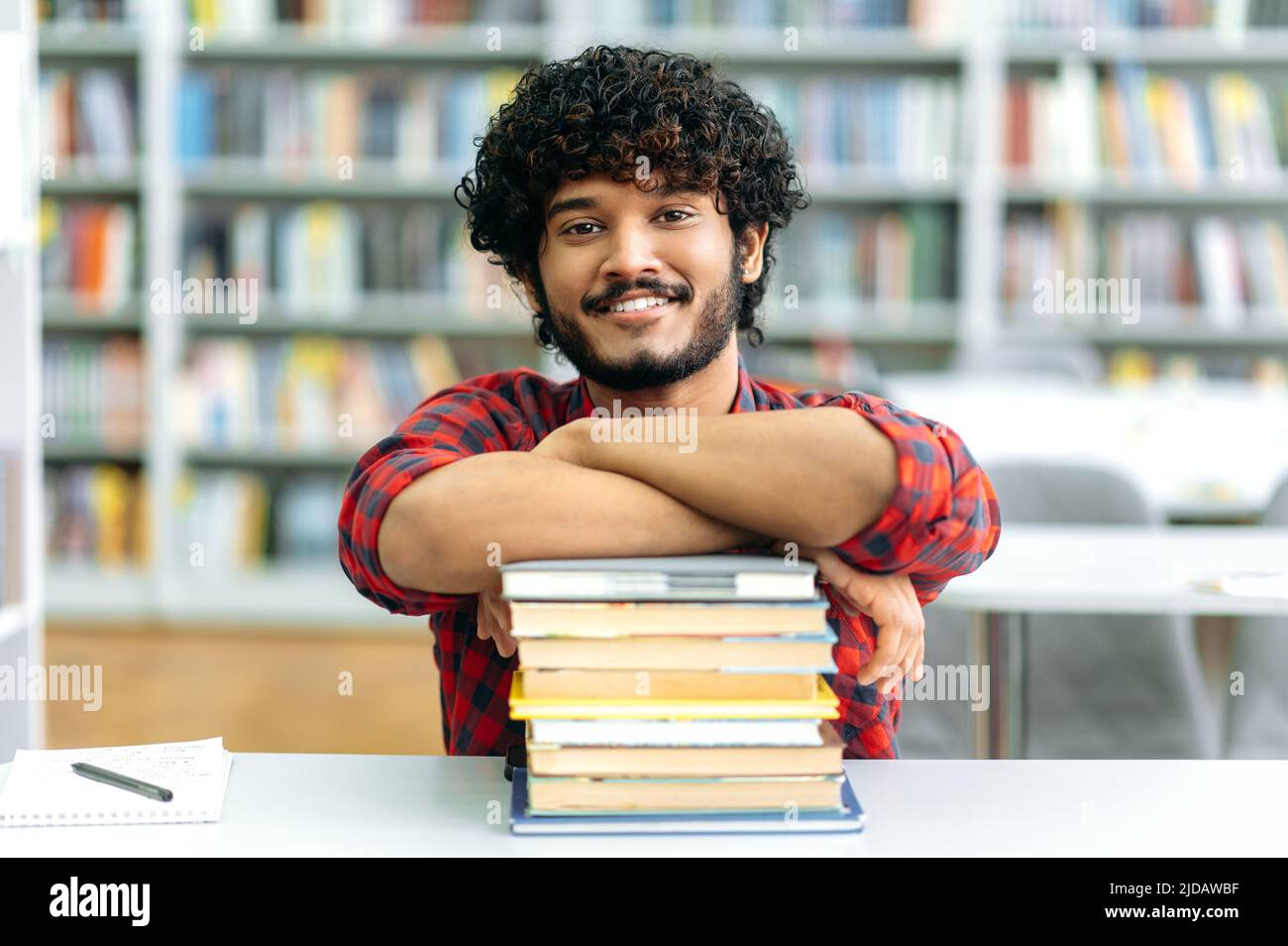 Positive clever indian or arabian guy, mixed race male, university student, sits at a desk with books, in the library against the background of bookshelves, looks at camera, smiles friendly Stock Photo