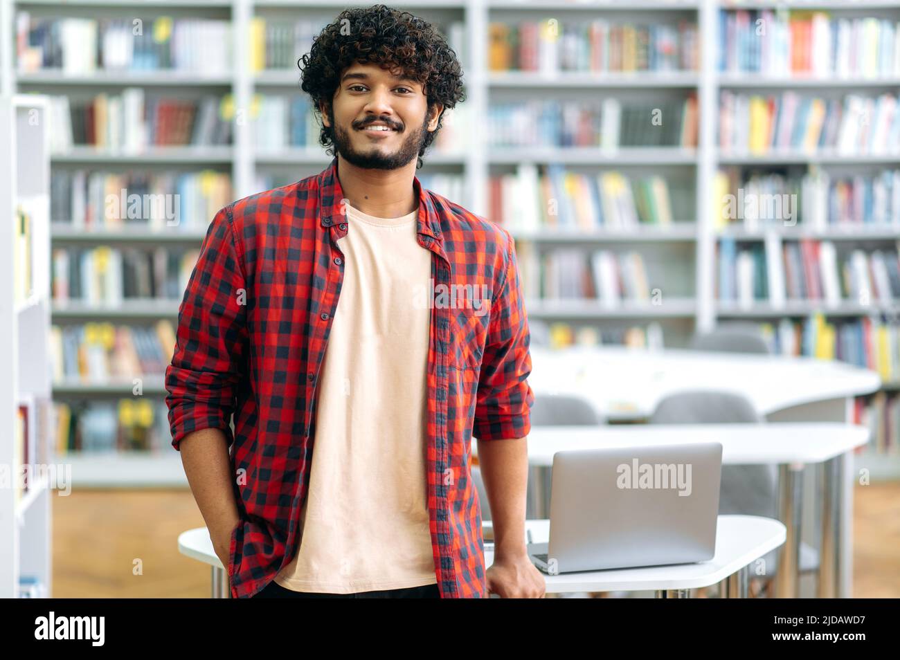 Handsome confident curly mixed race guy, Indian or Arabian male, in casual wear, student, freelancer or teacher, stands in the library against the background of bookshelves, looks at camera, smiles Stock Photo