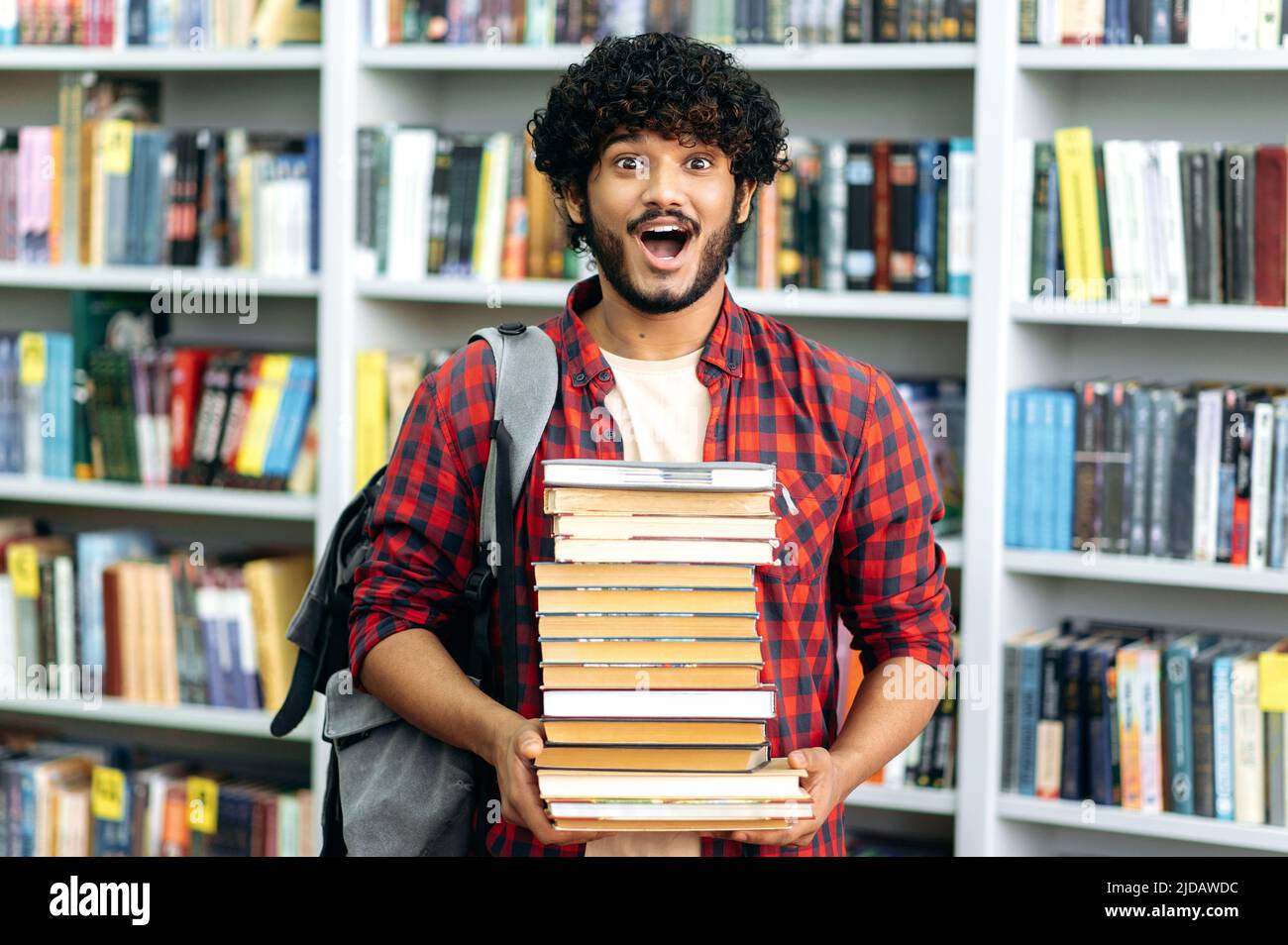 Excited happy amazed mixed race guy, university student, stands in the library against the background of bookshelves, holds a lot of books in his hands, looks at the camera in surprise, stunned Stock Photo