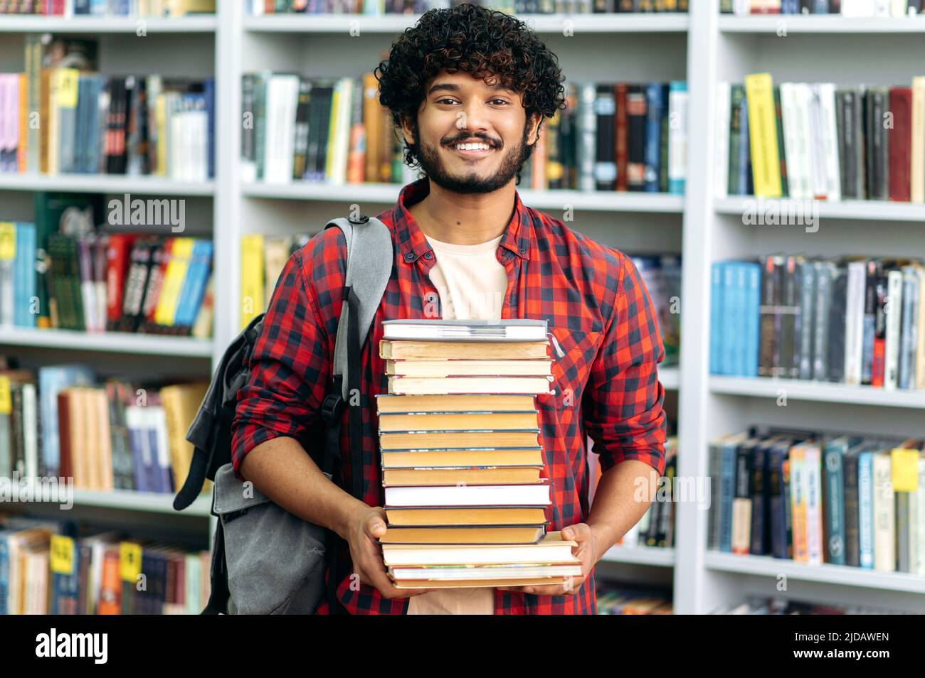 Happy smart indian or arabian guy, mixed race male, university student, stands in the library against the background of bookshelves, holds a lot of books in his hands, looks at camera, smiles friendly Stock Photo