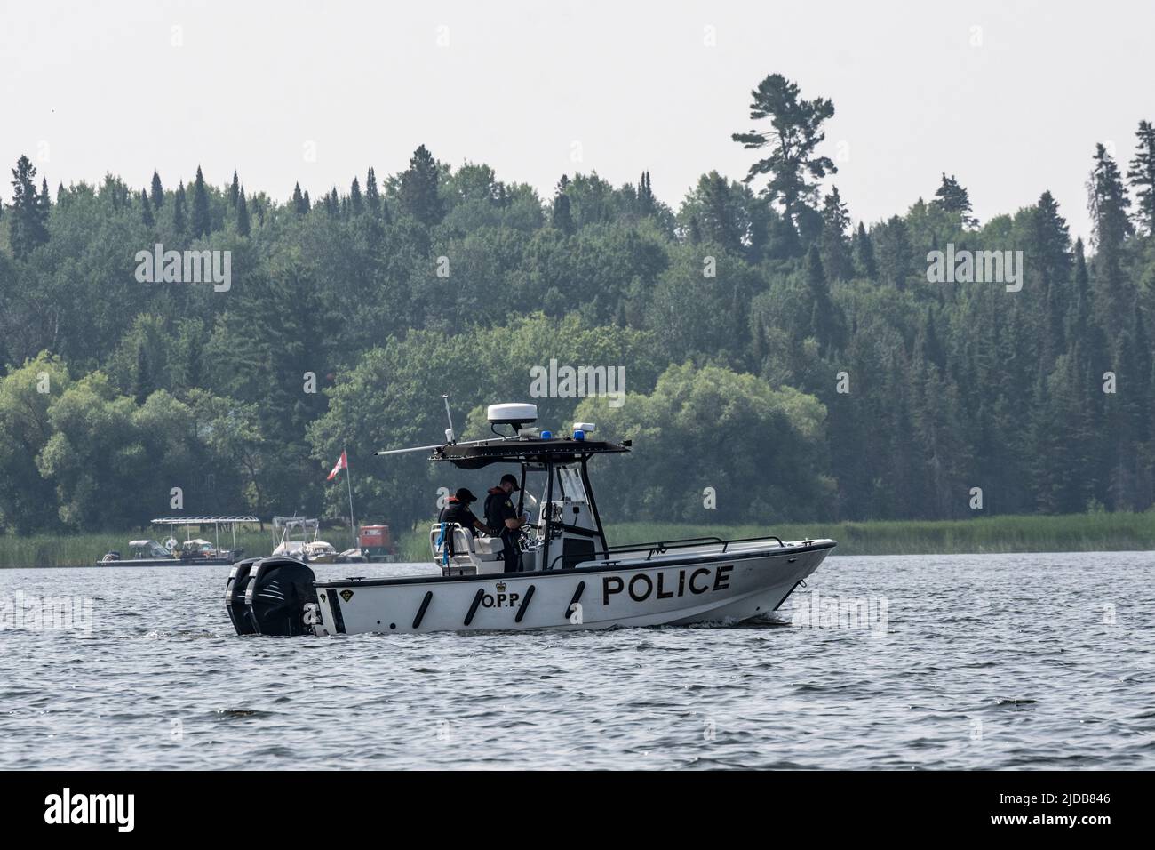 Police boat travelling along the shoreline of a lake past boats and docks, Lake of the Woods; Kenora, Ontario, Canada Stock Photo