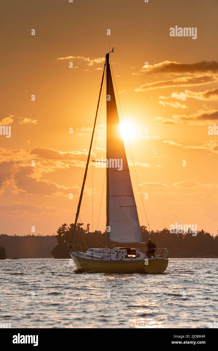Sailing on a lake at sunset with a golden sun glowing over silhouetted landscape and water, Lake of the Woods, Ontario; Kenora, Ontario, Canada Stock Photo