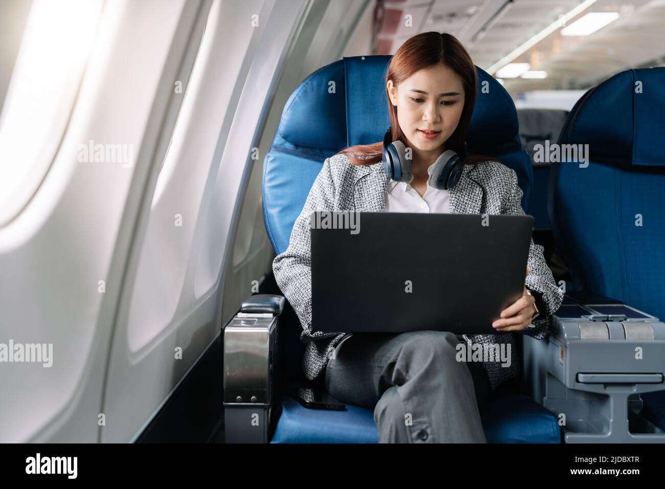Asian young woman wearing headphone using laptop sitting near windows at first class on airplane during flight, Traveling and Business concept Stock Photo