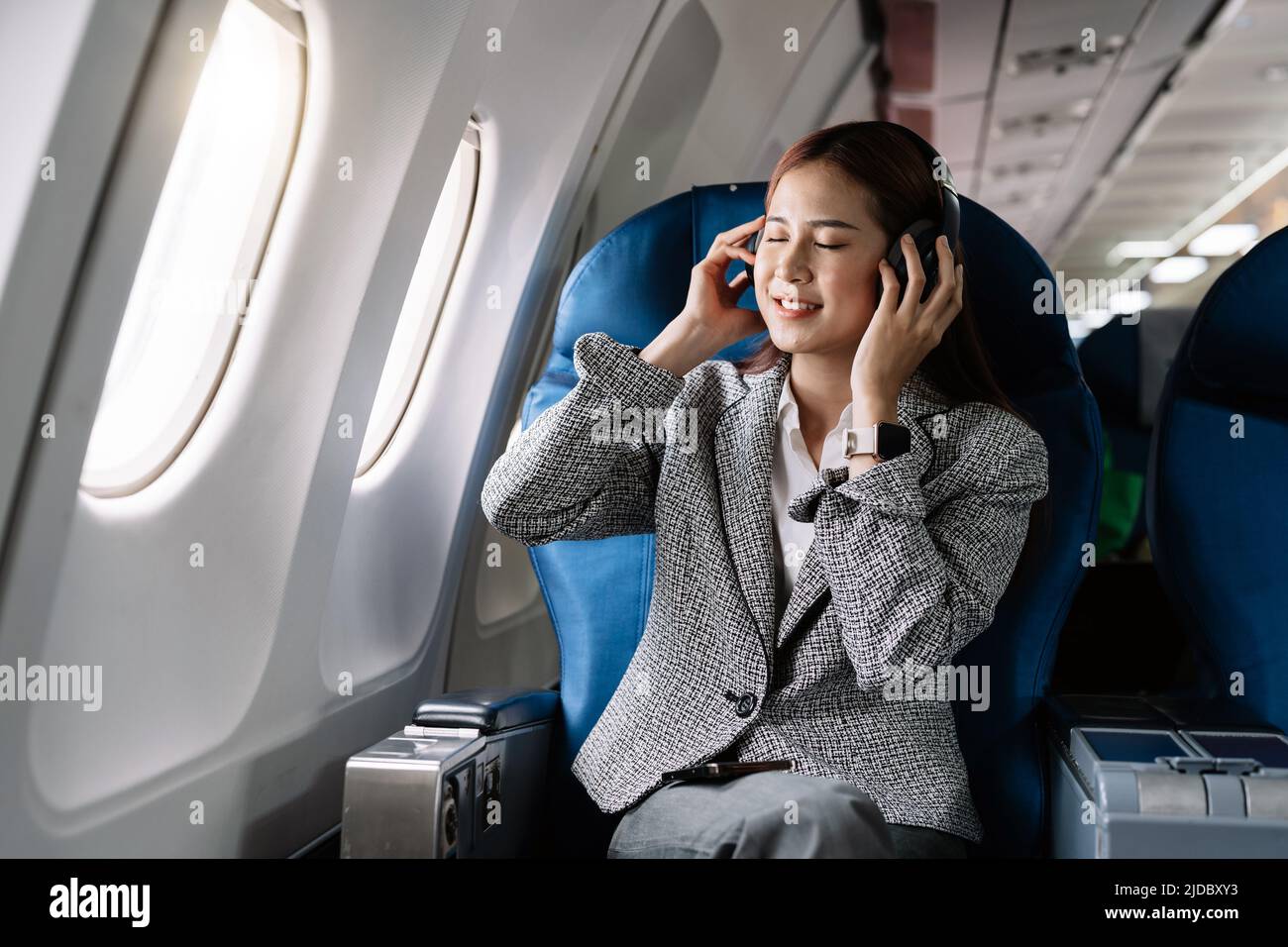 Young asian woman in wireless headphones listening to music and smiling during fly in airplane, travel concept Stock Photo