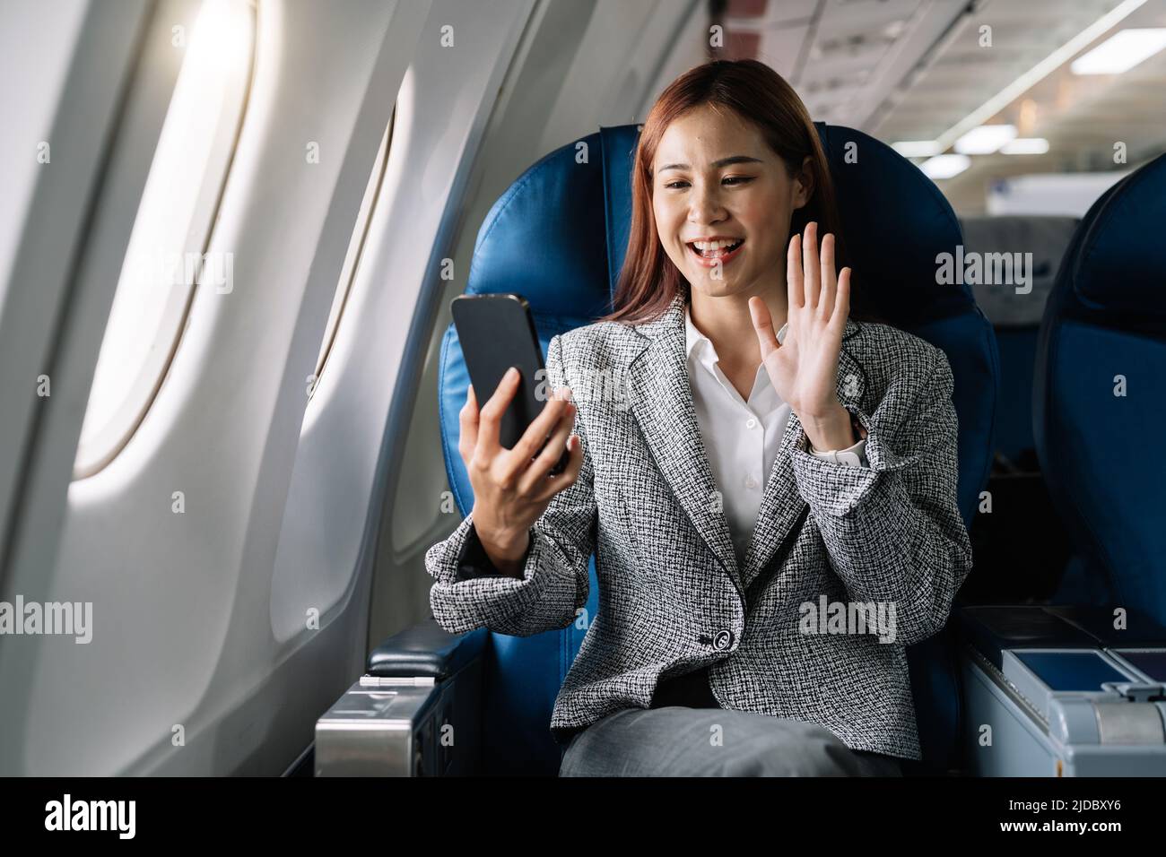 Smiling young asian businesswoman waving hand during video call on smartphone in plane Stock Photo