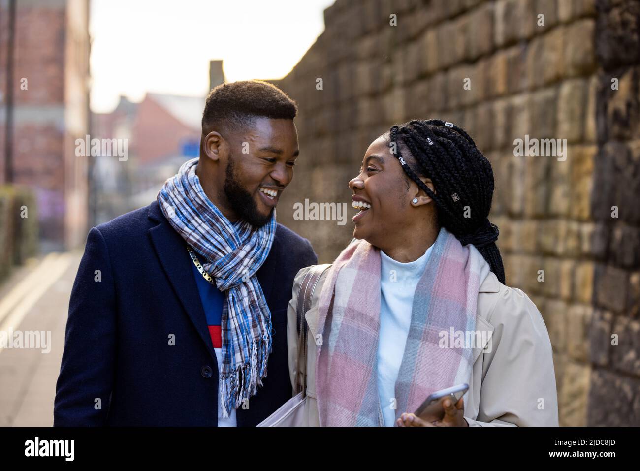 Cheerful couple wearing scarves chatting and walking past stone wall Stock Photo