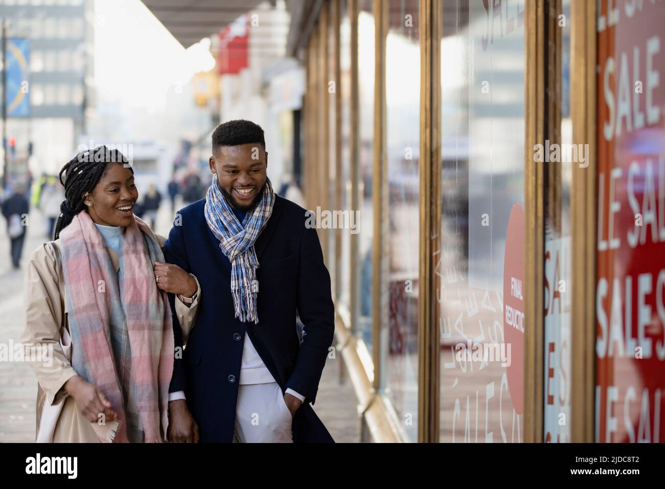 Cheerful couple arm in arms passing shop window in town and smiling Stock Photo