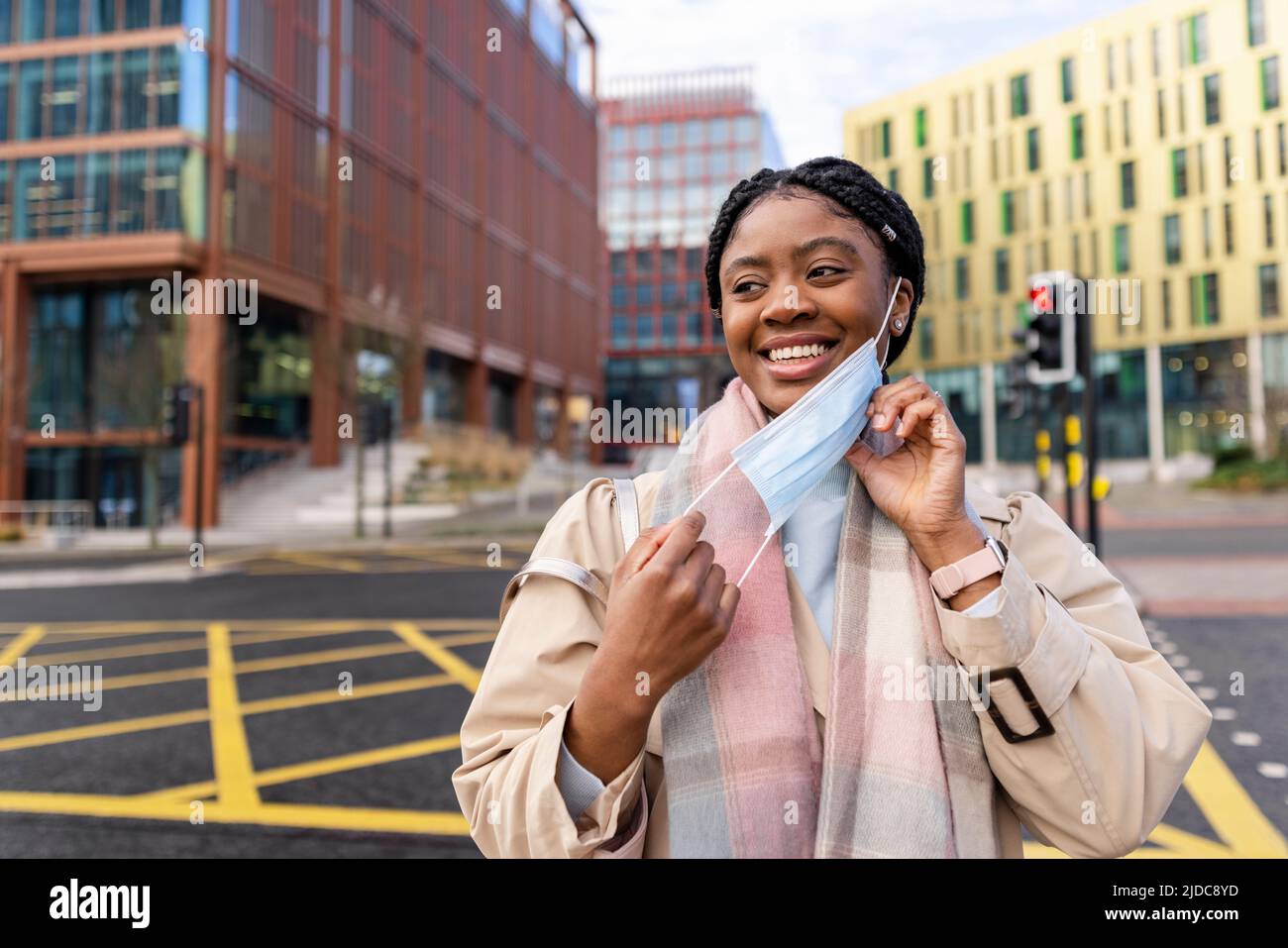 Cheerful woman in city centre taking off face covering, smiling and looking away Stock Photo