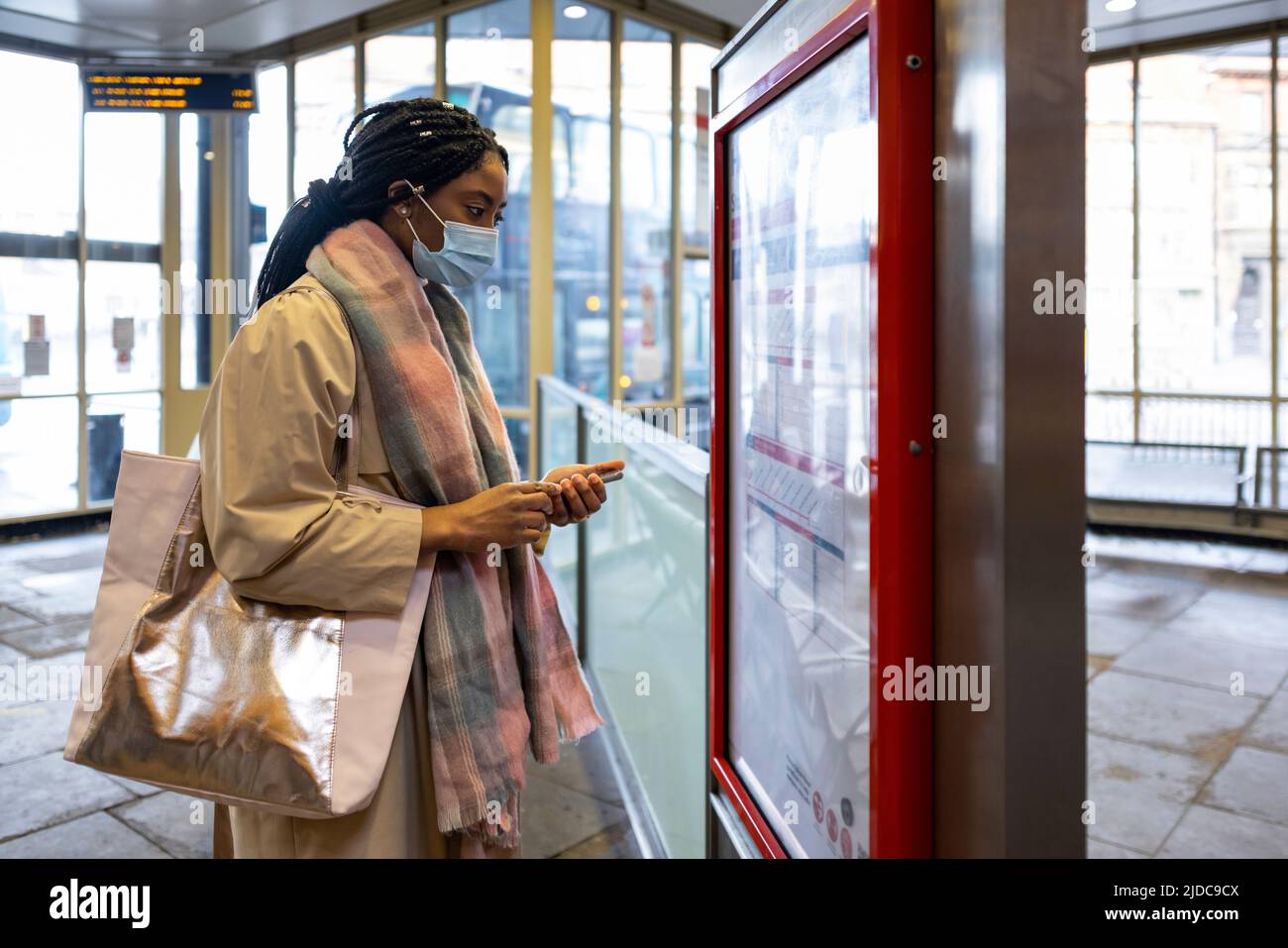 Woman with face covering holding phone and looking at bus timetable Stock Photo