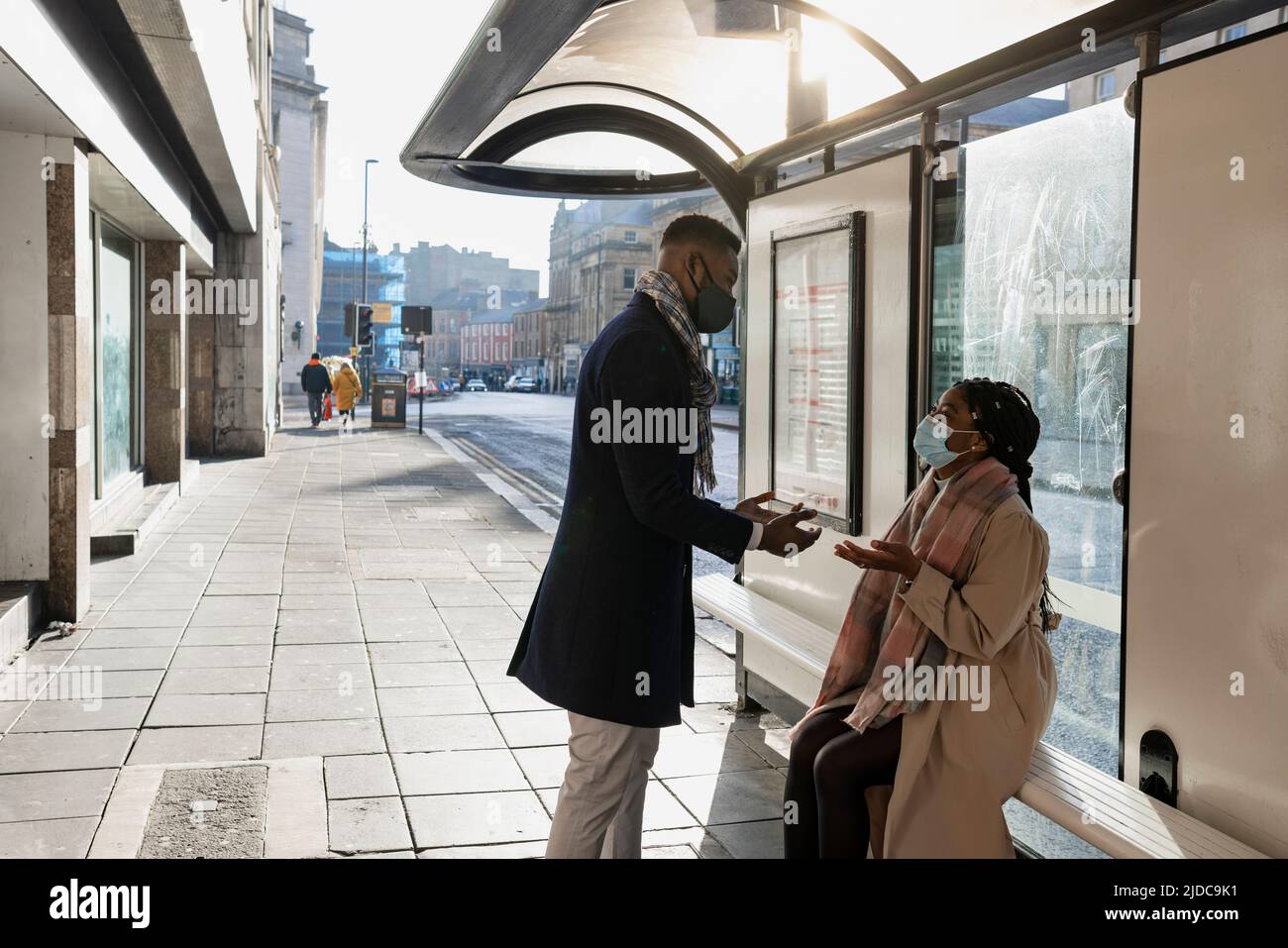 Two people wearing face coverings waiting for bus and having conversation, gesticulating with hands Stock Photo