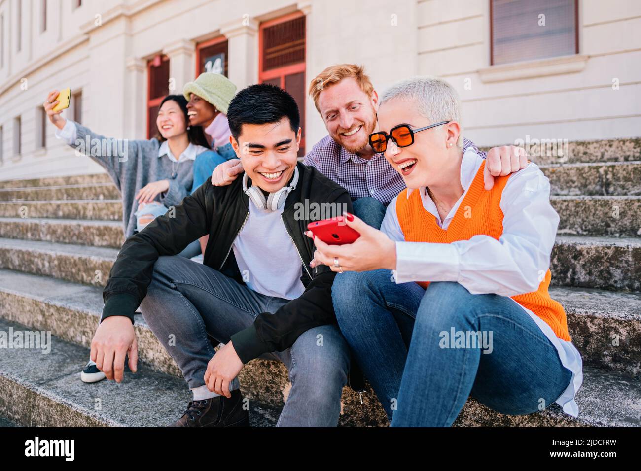 Group of young people sitting at outdoor watching socialmedia in the smartphone. Happy multiethnic hipsters looking funny online content on mobile Stock Photo