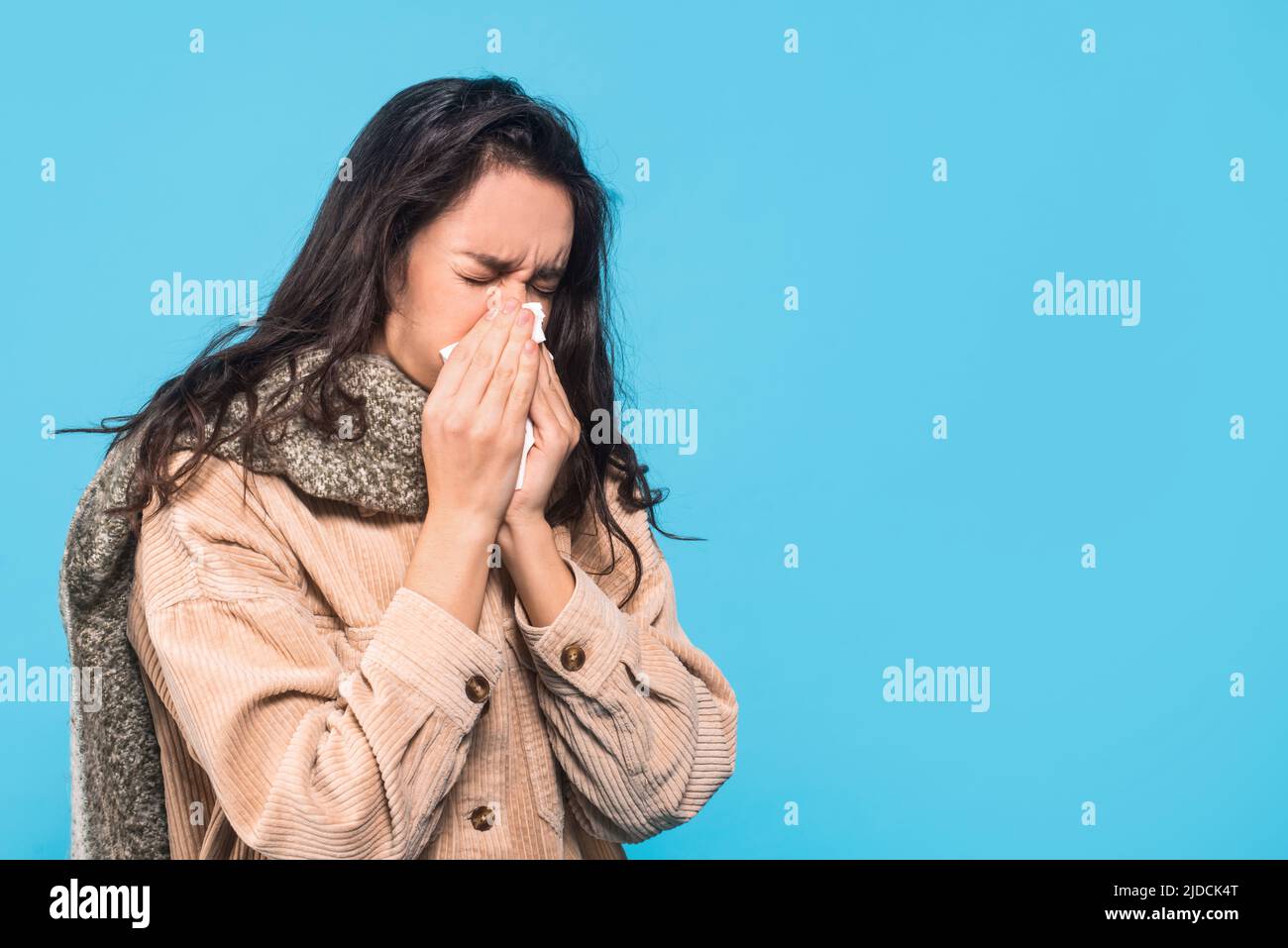 Sad millennial caucasian woman brunette patient with scarf blowing her nose, isolated on blue background Stock Photo