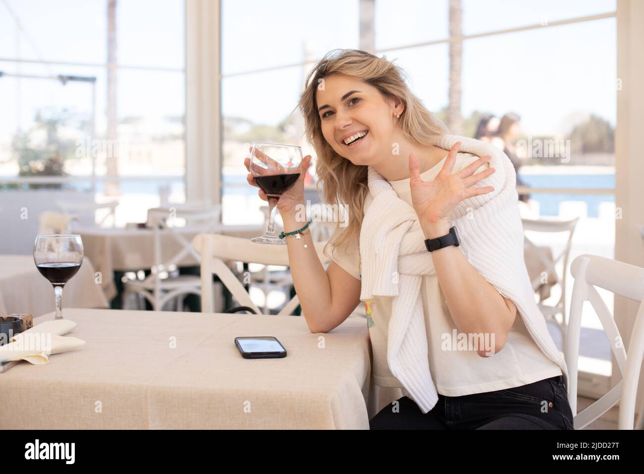 Portrait of young happy woman sitting at table near smartphone, holding glass of wine in cafe, spreading hands greeting. Stock Photo