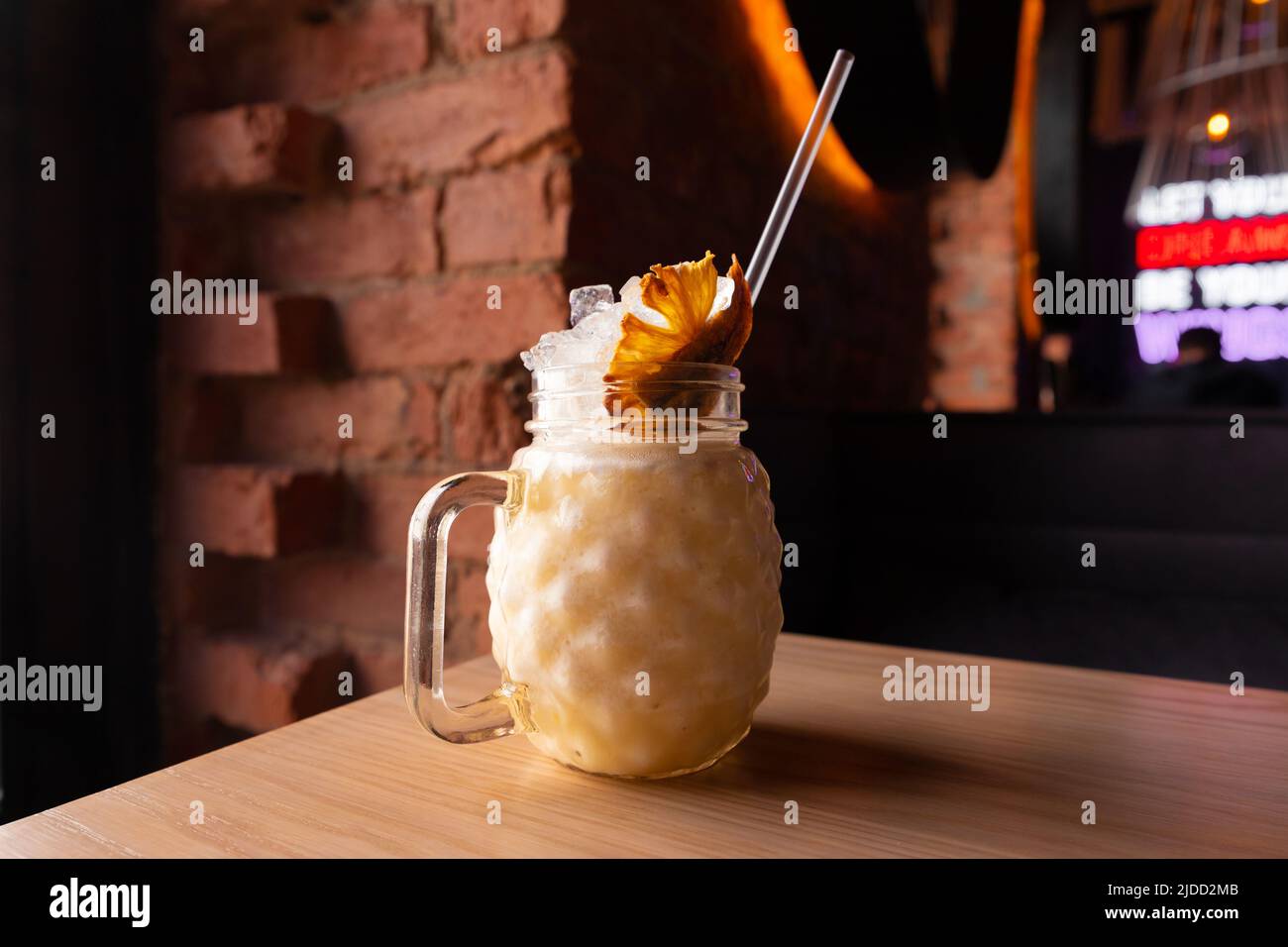 Cocktail Banana pina colada with ice, cocktail tube and pineapple chips in glass with handle on wooden table on brick background.  Bar or cafe. Front Stock Photo