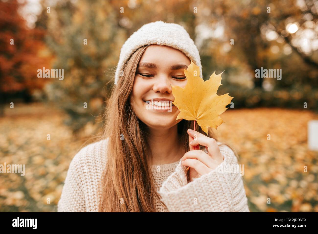 Charming young woman in knitted hat and sweater smiles happily on walk in autumn park, holding yellow maple leaf in front of her. Enjoying autumn Stock Photo