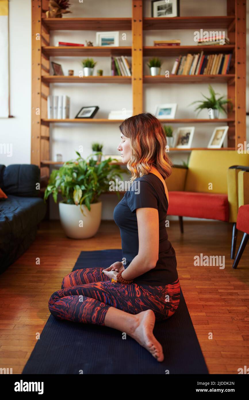 A middle-aged woman in yoga pants sits on a yoga mat at home in hero yoga pose and meditating. A yogi woman meditating at home. Stock Photo