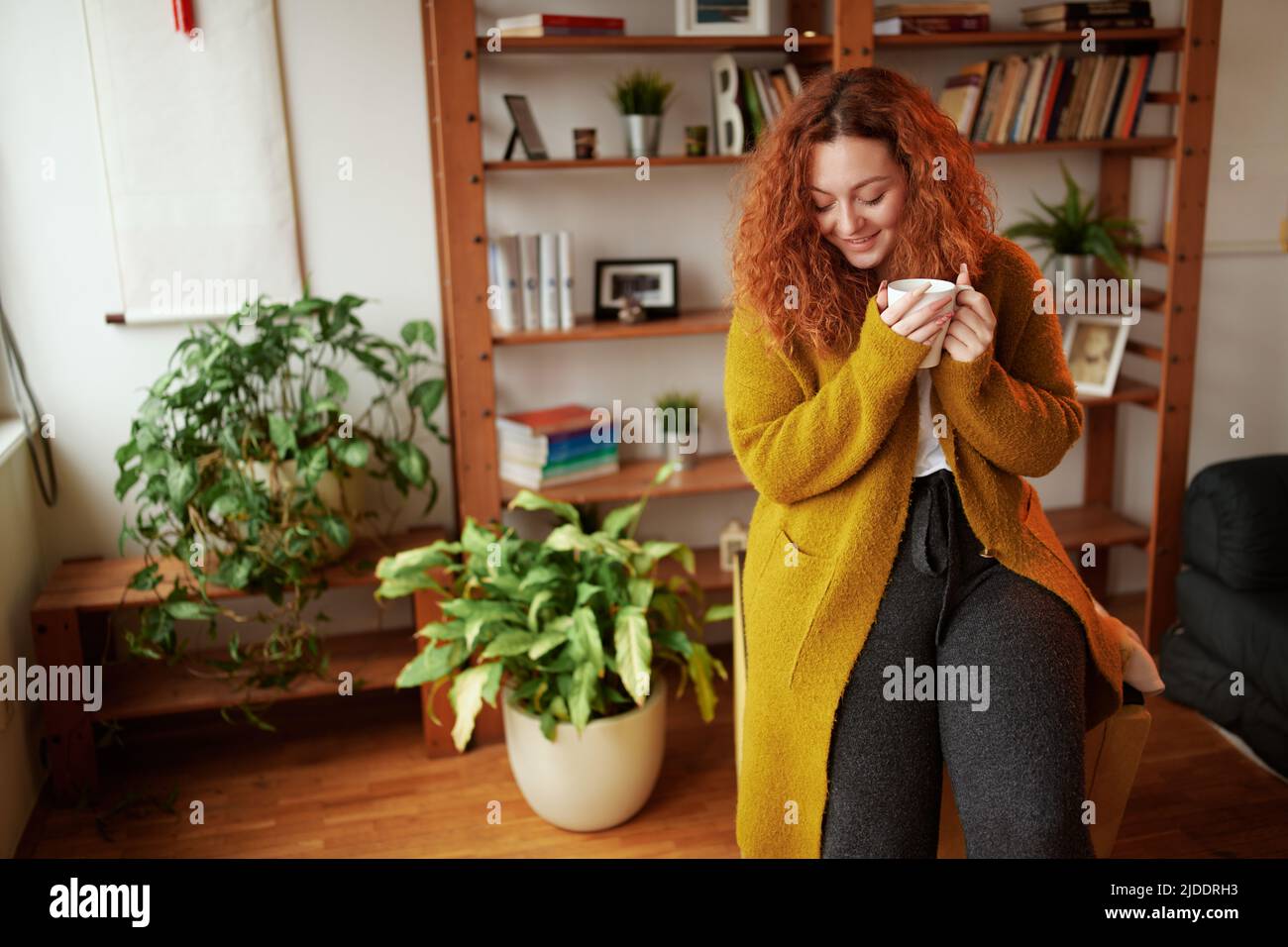 A happy young ginger girl is standing in the living room in the morning and enjoying her morning coffee. Stock Photo
